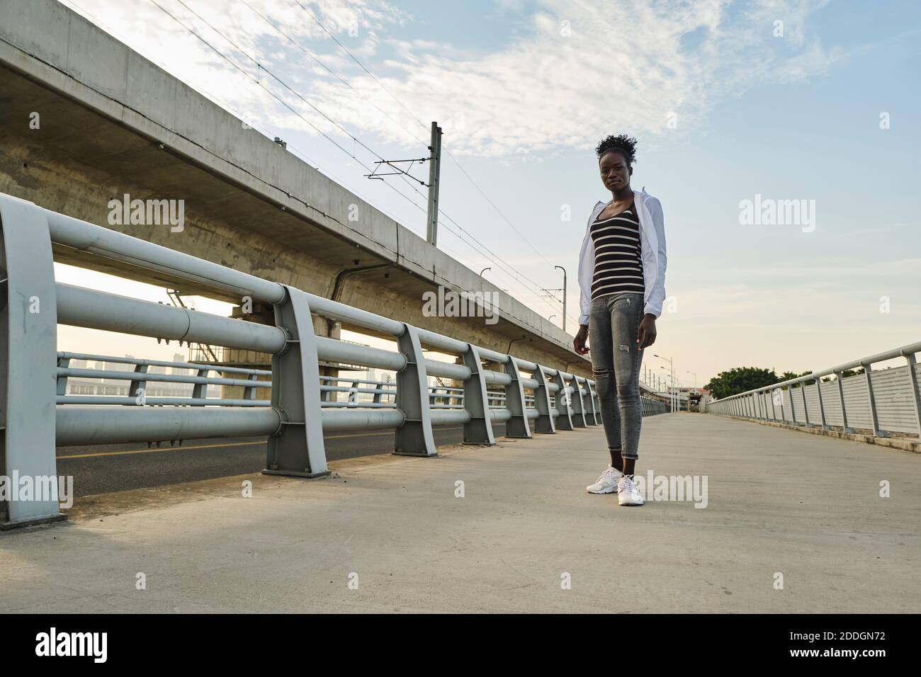 Corpo pieno di giovane studentessa nera sicura con Afro capelli che indossano abiti casual guardando la macchina fotografica mentre si sta in piedi vicino recinzione sulla città emba Foto Stock