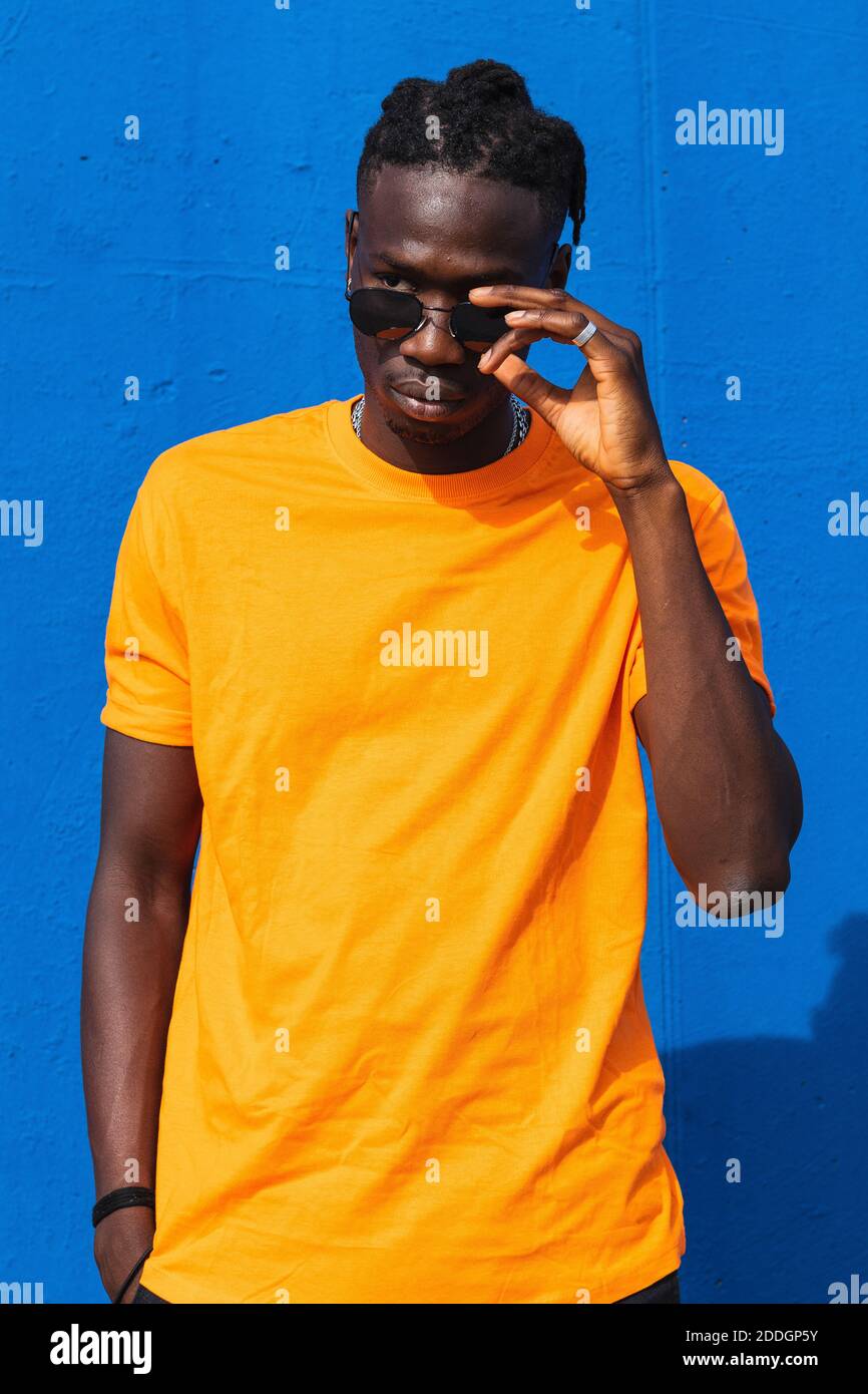 Serio giovane afroamericano ragazzo in maglietta gialla luminosa e occhiali da sole alla moda che guardano lontano su sfondo blu Foto Stock