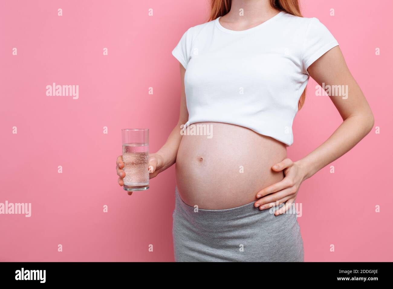 Ragazza incinta che tiene un bicchiere d'acqua sullo sfondo di un ventre incinta, su uno sfondo rosa, il concetto della quantità di acqua consumata Foto Stock