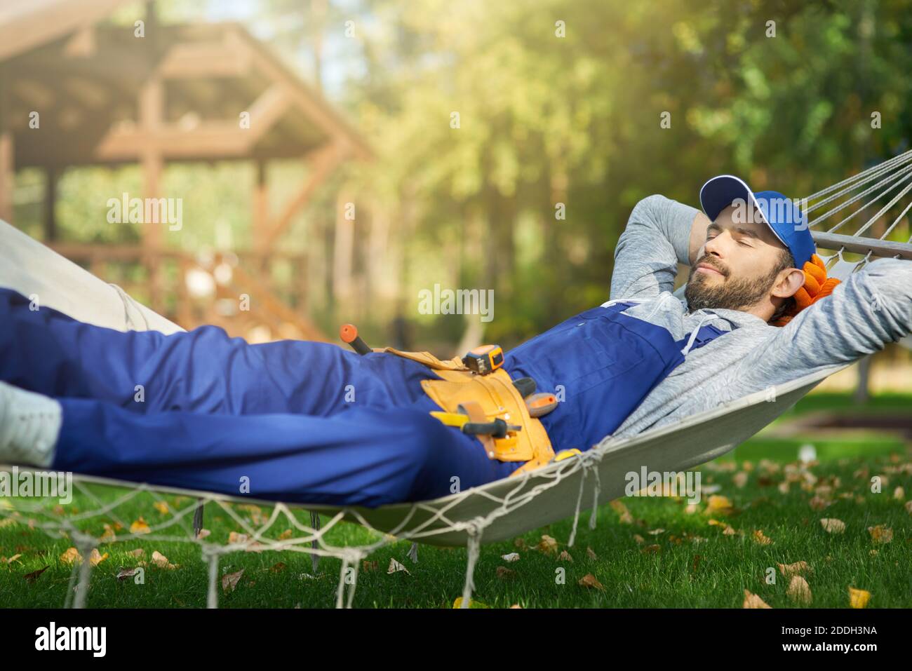 Rilassato giovane costruttore maschile indossando tute blu e berretto prendendo una pausa, sdraiato in un amaca all'aperto con gli occhi chiusi in una giornata di sole. Costruzione, professione, concetto di riposo Foto Stock