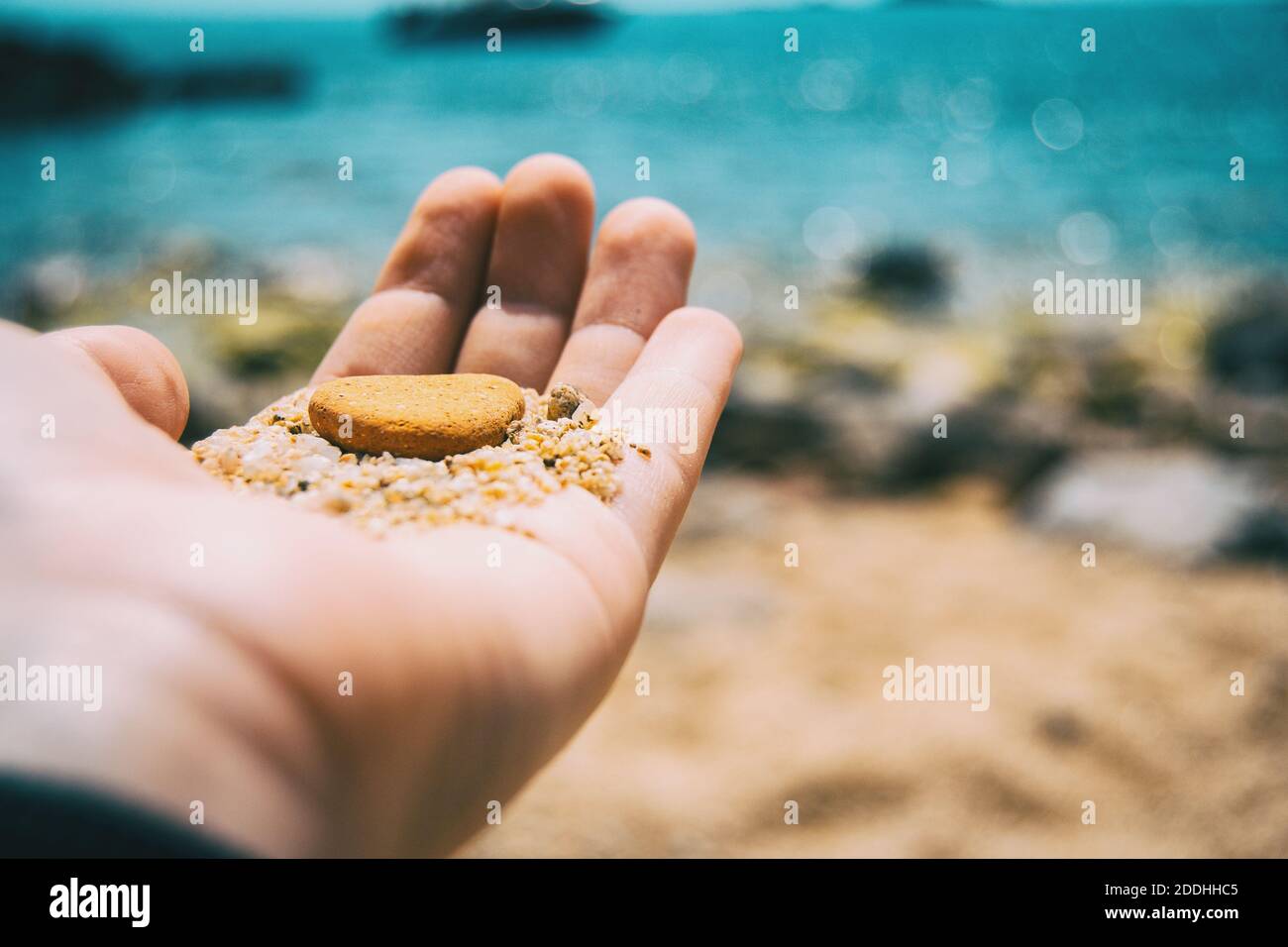 Primo piano di una mano che tiene su un ciottolo marrone e. una certa sabbia su uno sfondo non focalizzato del mare Foto Stock