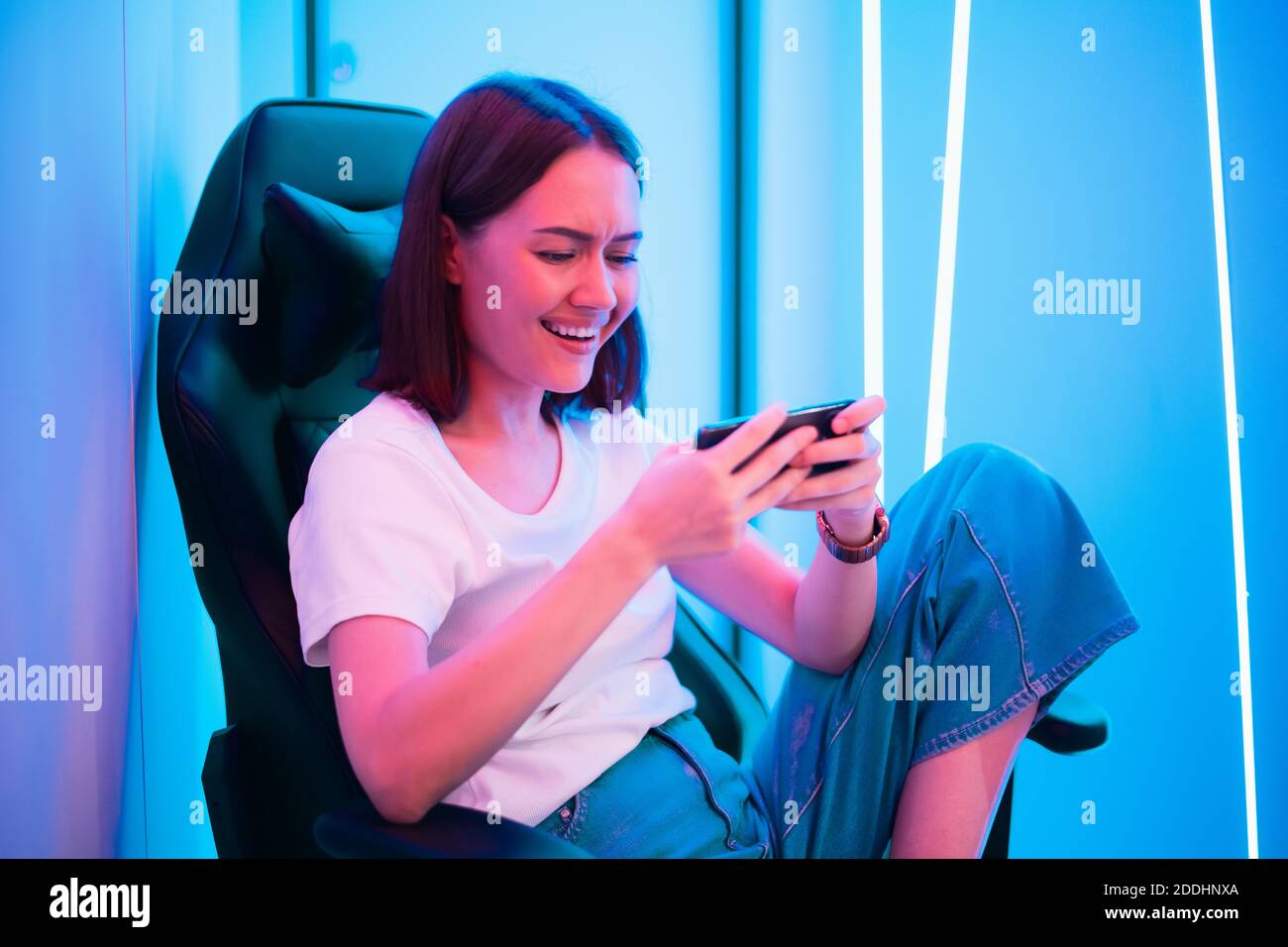 Carino ragazza giocatore sedersi su una sedia di gioco, sorridente e giocare gioco mobile online su uno smartphone. Divertirsi giocando con gli amici online. Foto Stock