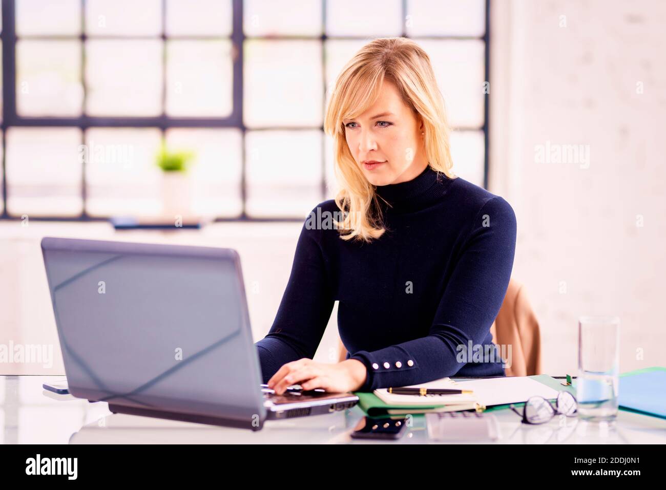 Foto di una donna d'affari bionda sorridente seduta alla scrivania dietro il suo laptop e funzionante. Ufficio domestico. Foto Stock