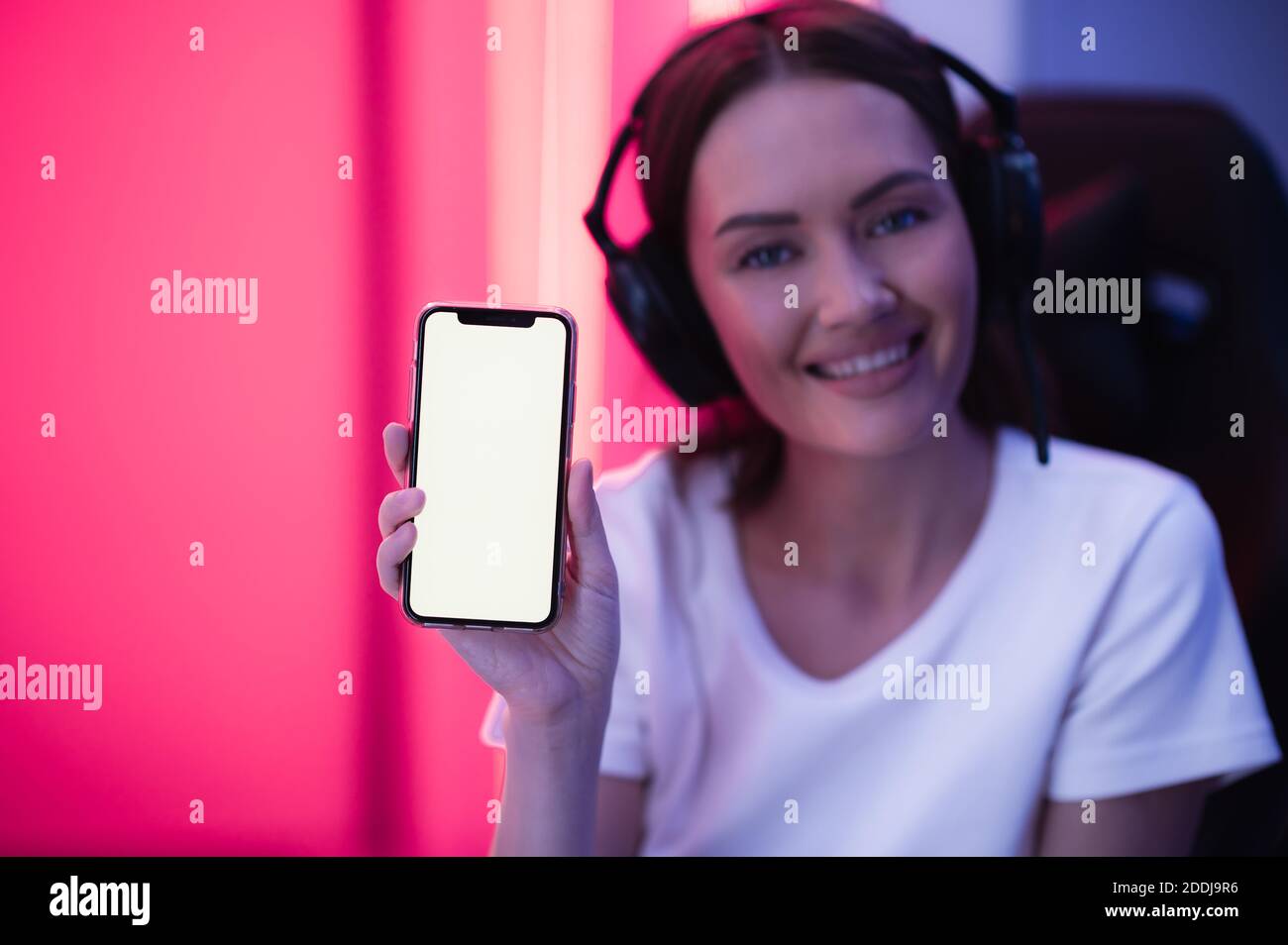 Ragazza felice del giocatore che indossa la cuffia che mostra il display dello smartphone e punta verso lo schermo vuoto con il dito. Foto Stock