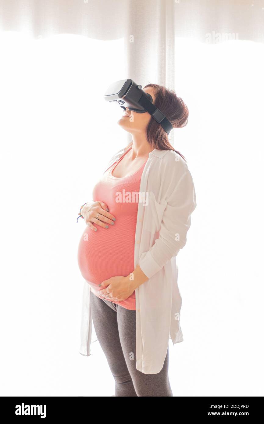 Giovane bella donna incinta che indossa occhiali di realtà virtuale a casa. Concetto di connessione, tecnologia, nuova generazione e progresso Foto Stock