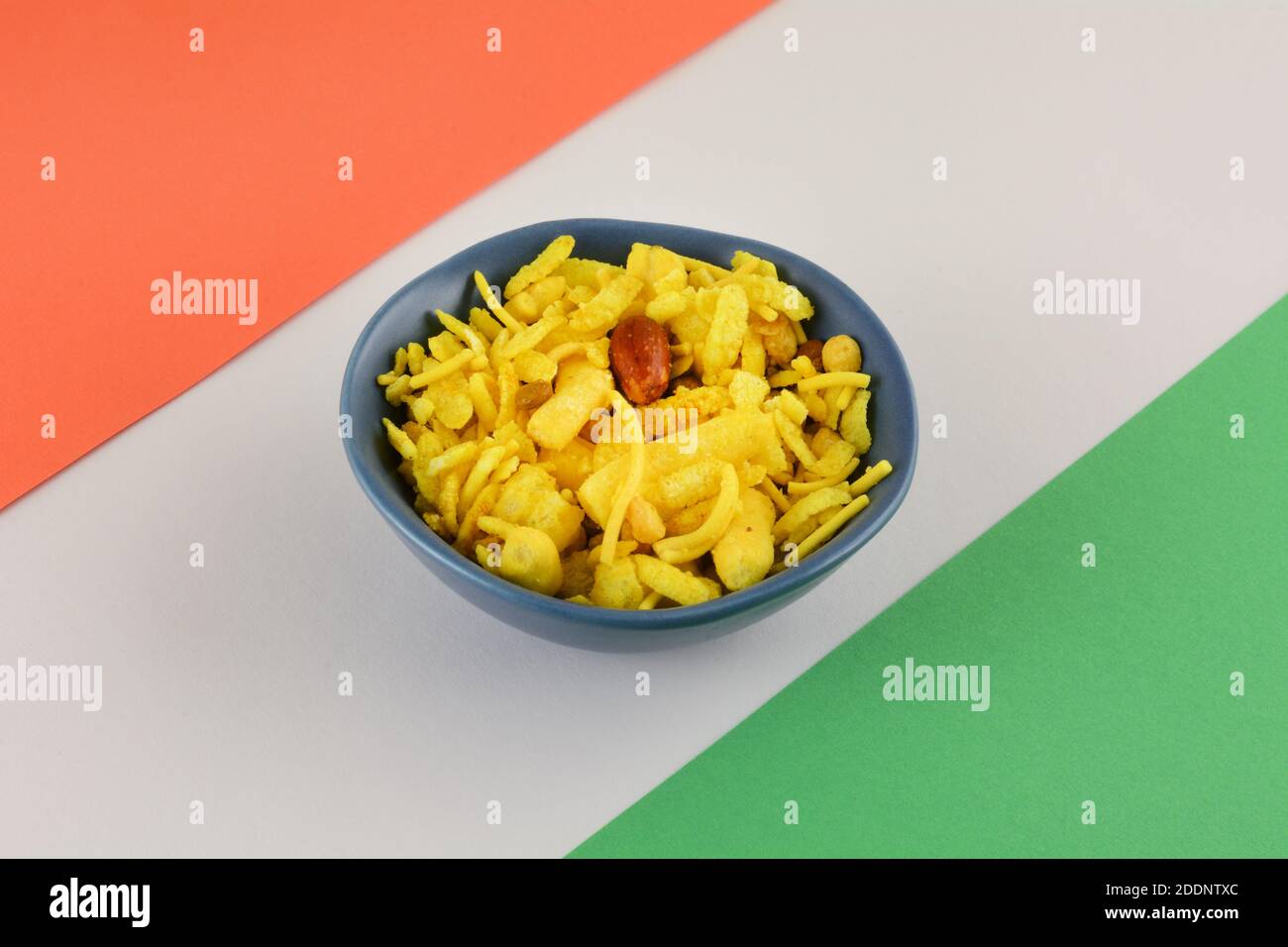 Spuntino indiano dolce e aspero di tagliatelle, arachidi, riso soffiato e piselli in una ciotola blu su fondo a strisce arancione, bianco e verde Foto Stock
