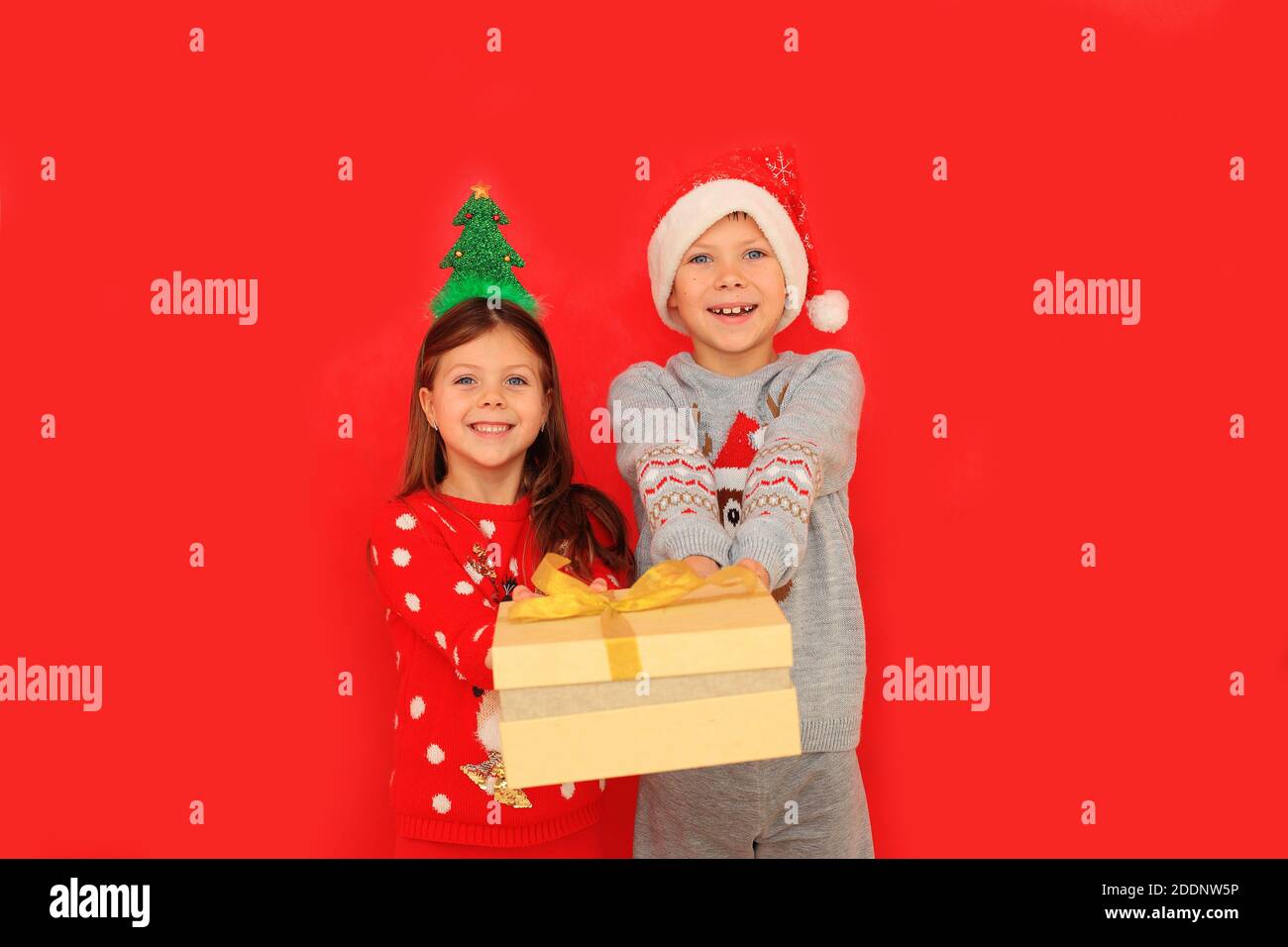 Due bambini in pullover hanno in mano un regalo di Capodanno su sfondo rosso. Un ragazzo e una ragazza danno un regalo di Natale a vicenda Foto Stock