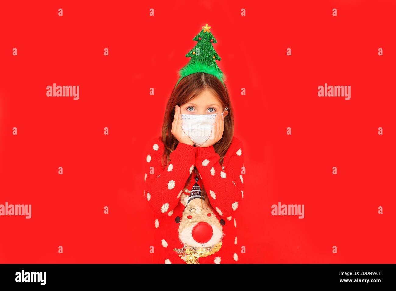 Bambino in una maschera respiratoria e un maglione di Capodanno su sfondo rosso. Ragazza in un pullover rosso di Natale durante il periodo di quarantena Foto Stock
