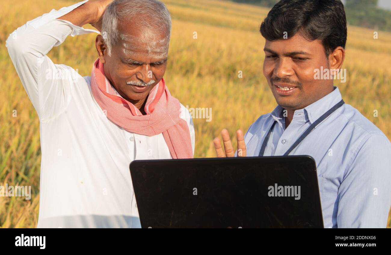 Confuso agricoltore indiano graffiare la testa mentre banchiere o ufficiale del governo aziendale discutere sul portatile circa rendimento del raccolto, credito e prestito sovvenzione Foto Stock