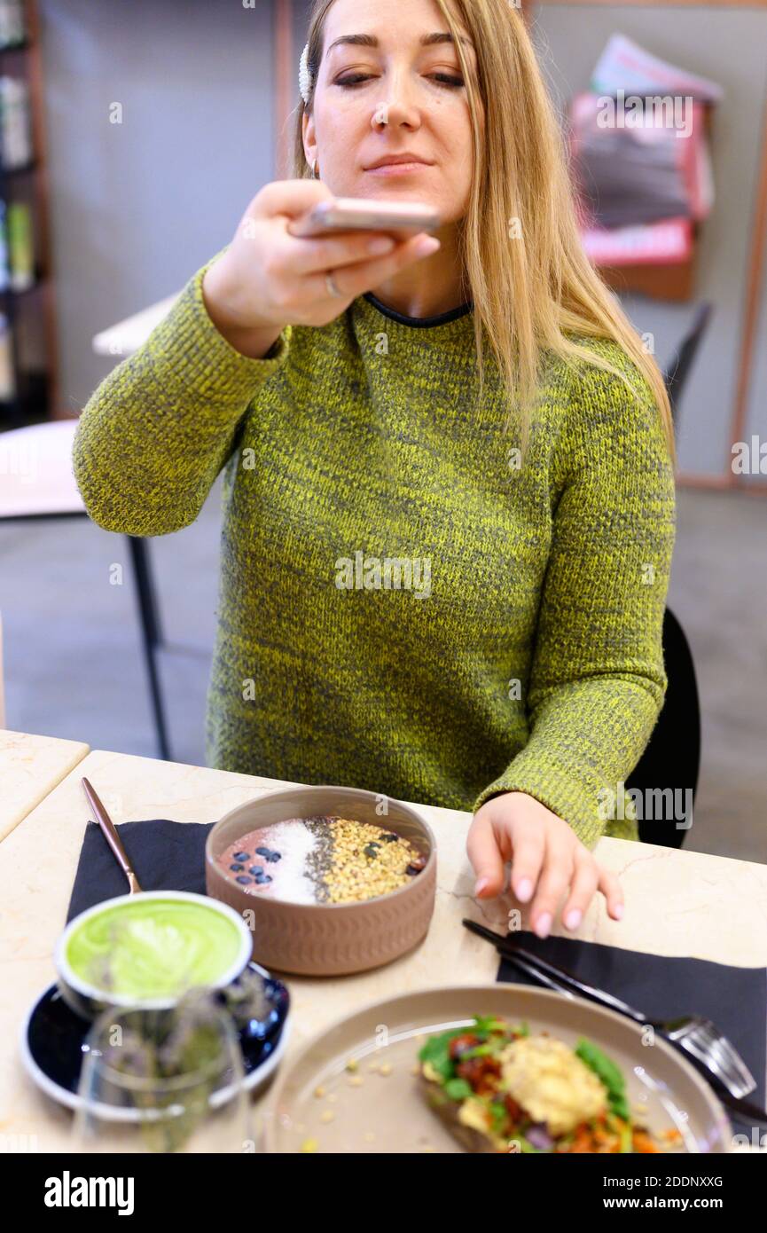 una donna scatta delle foto con il telefono del suo cibo crudo in un caffè. una ciotola frullato e una tazza di tè verde latte matcha e brinda con hummus e verdure cotte al forno Foto Stock