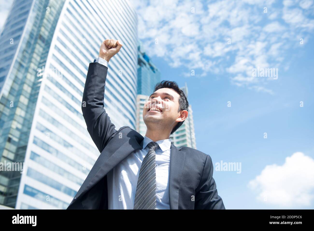 Imprenditore sorridente e sollevando il pugno in aria, con edificio per uffici in background - il successo aziendale, conseguimento e vincere dei concetti Foto Stock