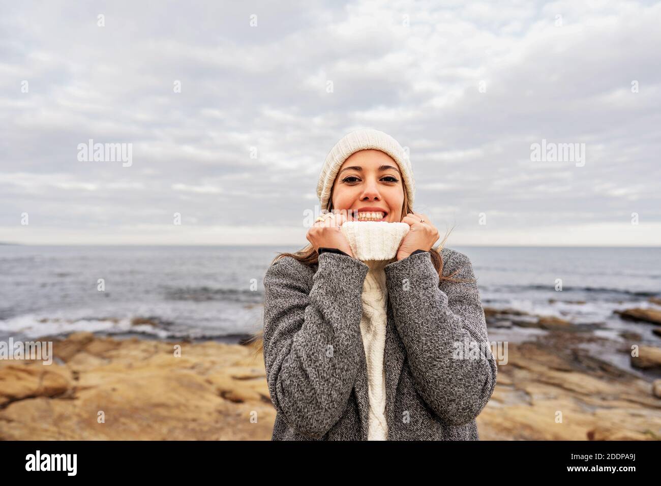 Bella giovane donna caucasica stringe il collo del suo bianco maglione con le mani per il freddo inverno sorridente guardando la macchina fotografica sul mare Foto Stock