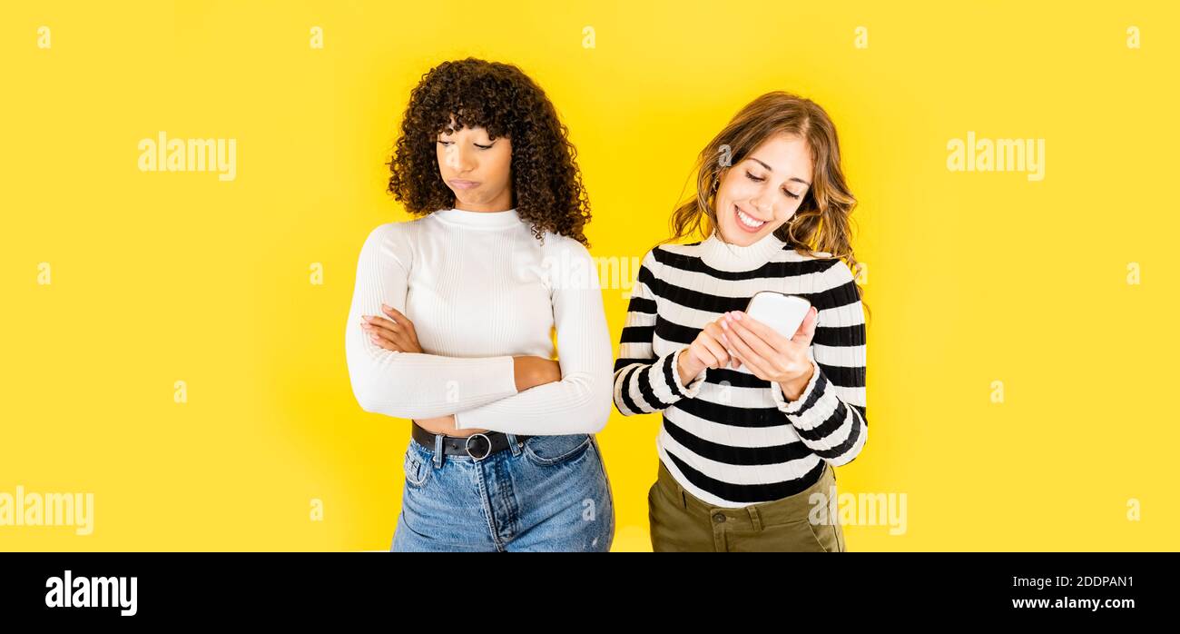 Due giovani donne di razza mista isolato divertente immagine sul giallo Sfondo per la copia spazio - donna caucasica che si diverte con Smartphone mentre il suo nero Foto Stock