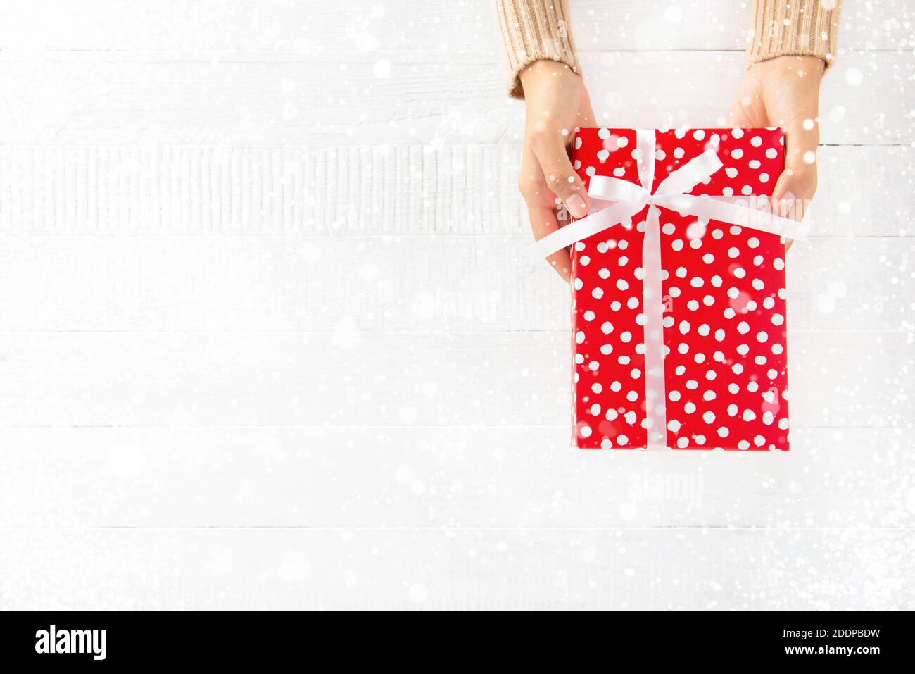 Mani donna che danno scatola regalo rossa su sfondo di legno bianco con nevicata Foto Stock