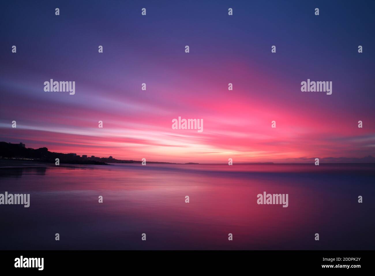 Una lunga esposizione cattura un cielo pieno di nuvole striate rosa e viola mentre il sole comincia a sorgere sul mare. La sabbia bagnata riflette i colori vivaci. Foto Stock