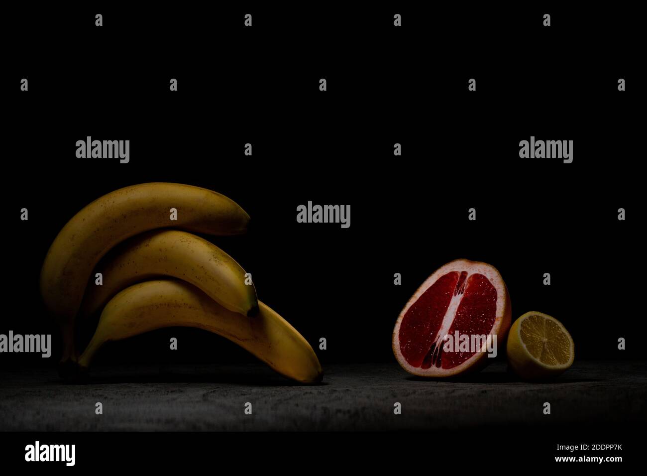 Tre banane mature giacciono l'una sull'altra su cemento grigio scuro, di fronte al limone e al pompelmo. Tasto basso. Foto Stock
