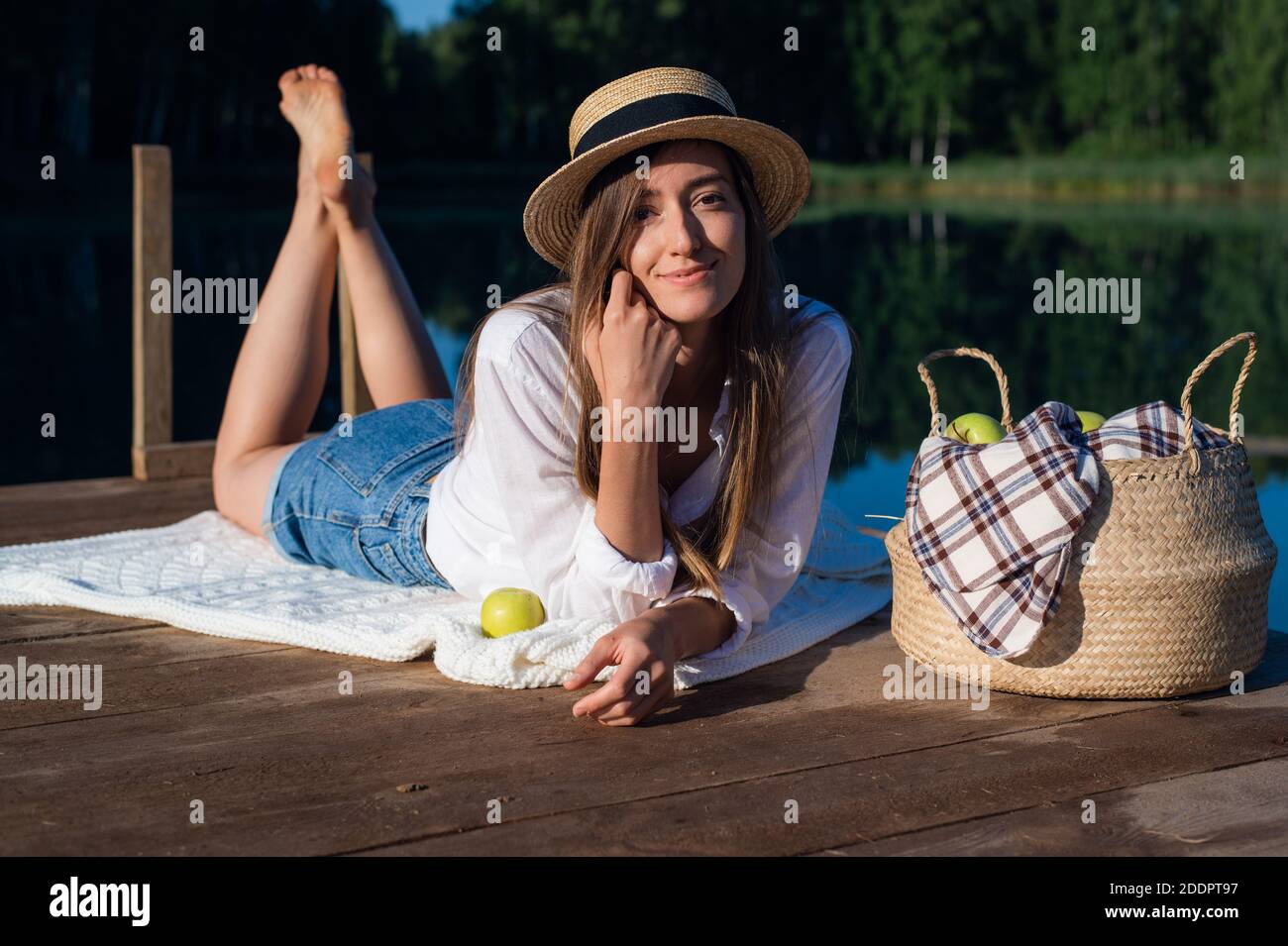 Giovane bella donna su un pic-nic presso il lago, sdraiata su un molo di legno. Indossa una camicia e un paio di pantaloncini in denim. Nelle vicinanze troverete un cesto di frutta e una mela. Foto Stock