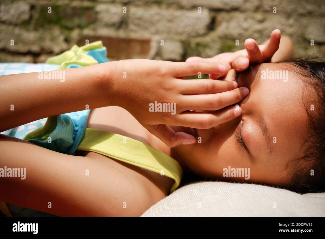 Etnia sudorientale bambina sdraiata su una panchina, prendere il sole la mattina a casa. Mani che vengono messe in faccia a coprire dalla luce solare dura Foto Stock