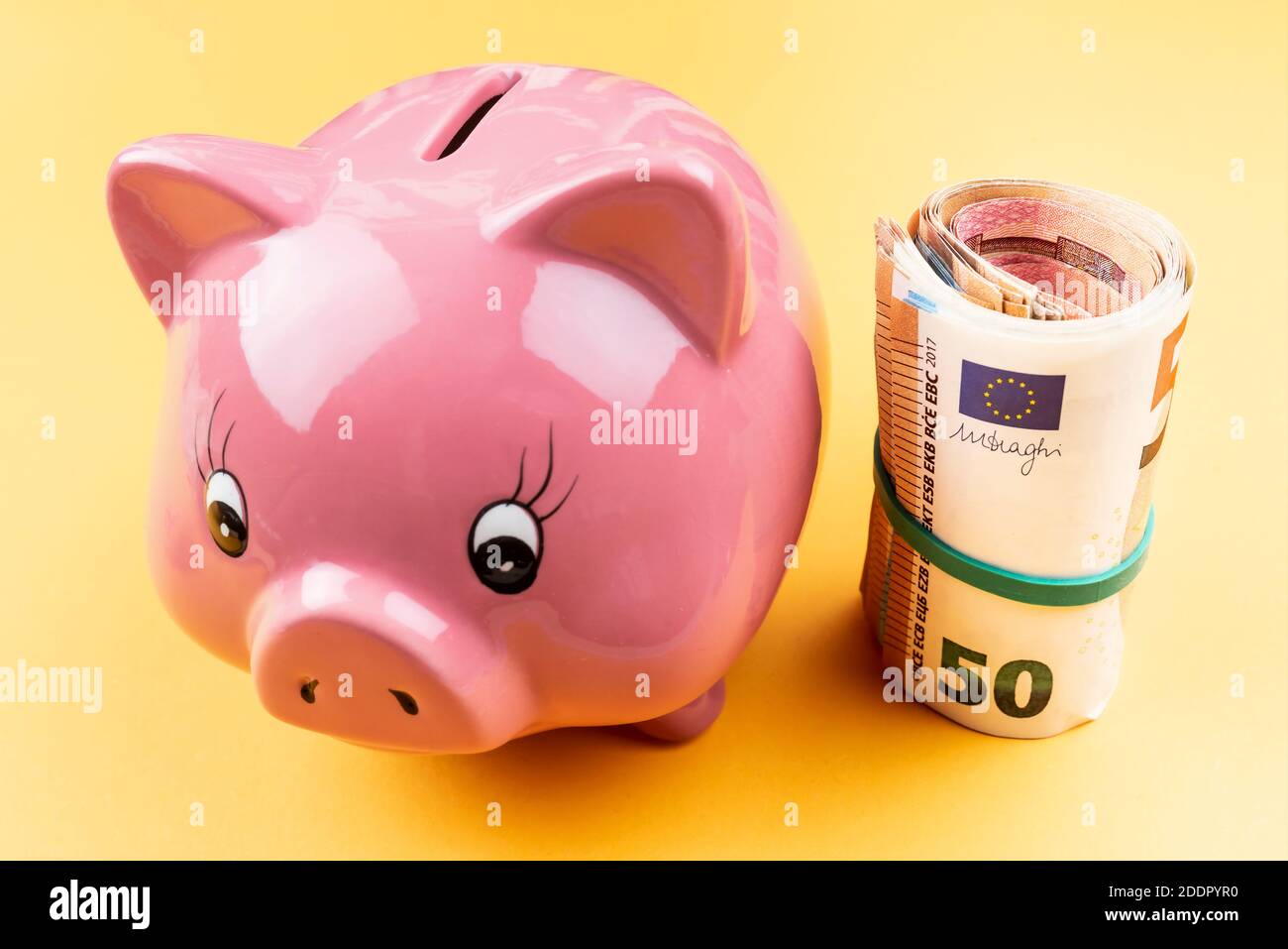vista ad alto angolo del rotolo di denaro con fascia in gomma accanto al banco di piggy rosa su sfondo arancione, concetto di risparmio Foto Stock
