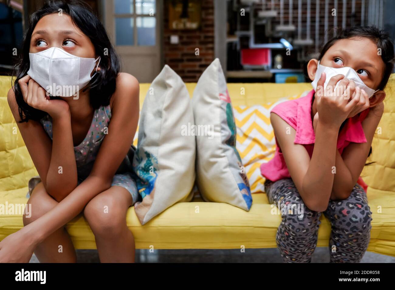 Due ragazze sudorientali piccole che indossano la maschera medica del viso ottenere Annoiato seduto su divano a parte l'uno dall'altro a casa Durante la pandemia Foto Stock
