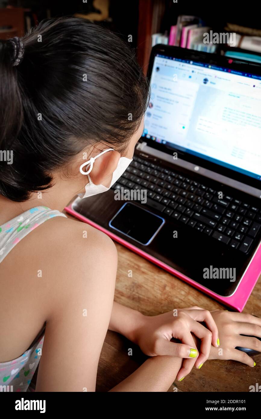 Ragazza teenage del sud-est asiatico con maschera facciale davanti al portatile a casa. Chat online con gli amici. Primo piano sul viso Foto Stock