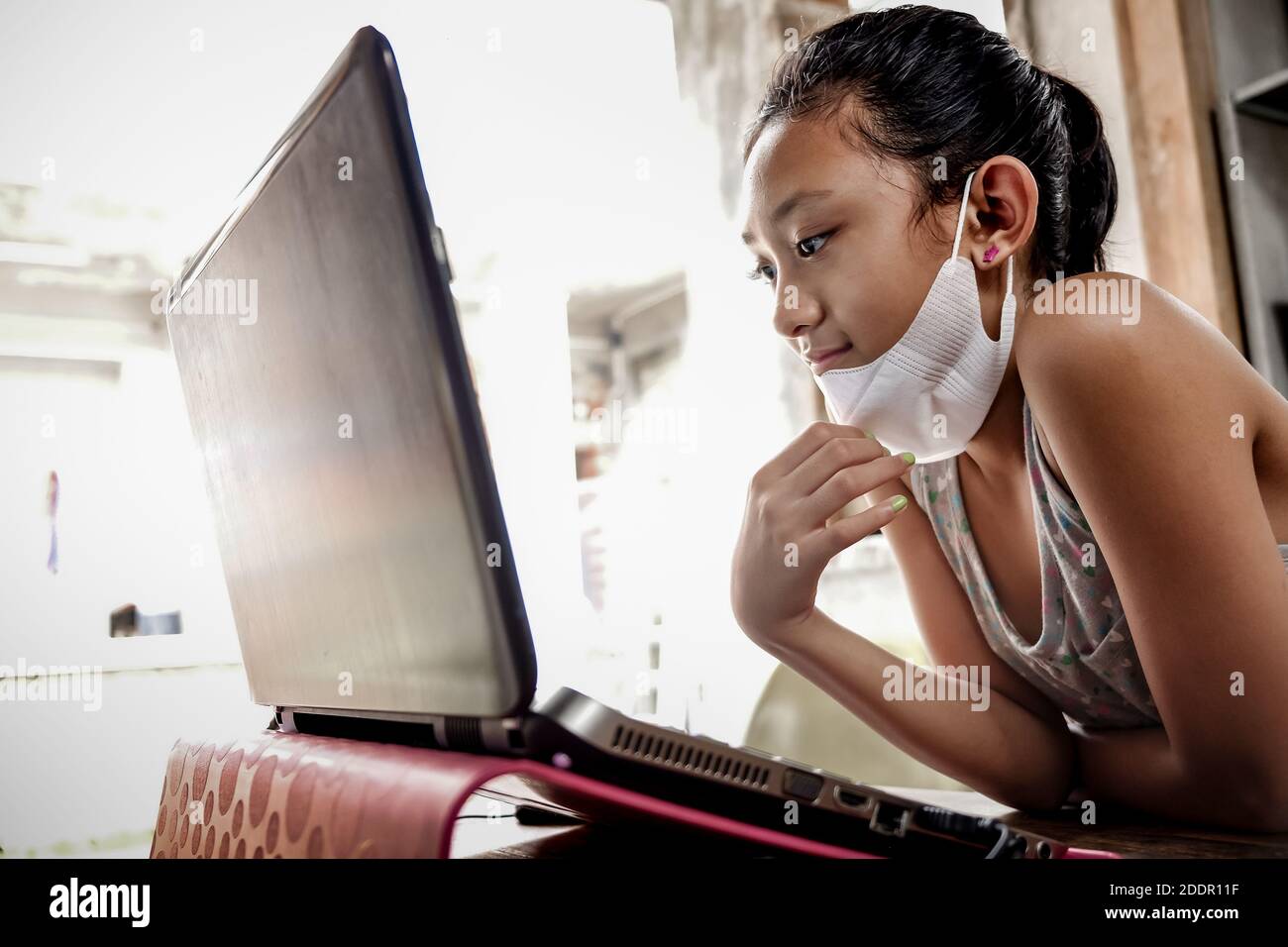 Ragazza teenage del Sud-est asiatico con maschera facciale davanti a un laptop a casa. Incontro online con gli amici durante la pandemia globale Foto Stock