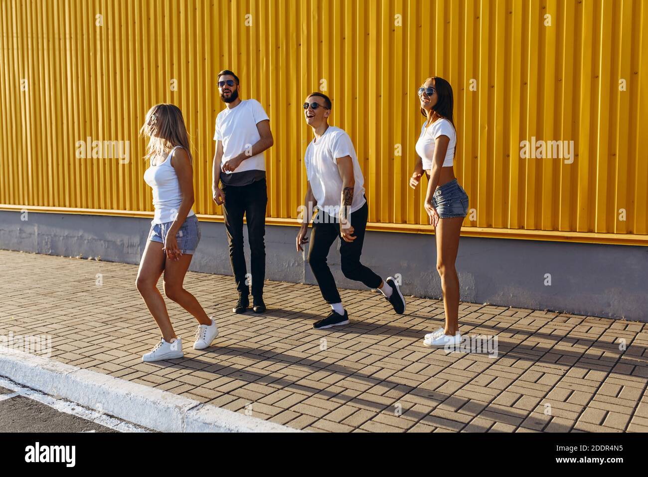 Felici giovani amici, uomini e donne, si divertono e saltano in giro per la città sullo sfondo del muro giallo, raffigurante un gesto di vittoria Foto Stock