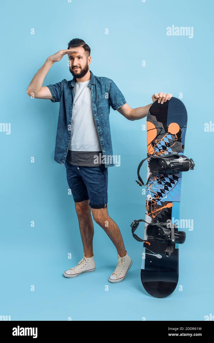 Un uomo felice, vestito di pantaloncini e camicia , tiene uno snowboard e guarda in lontananza, in piedi su uno sfondo blu isolato Foto Stock