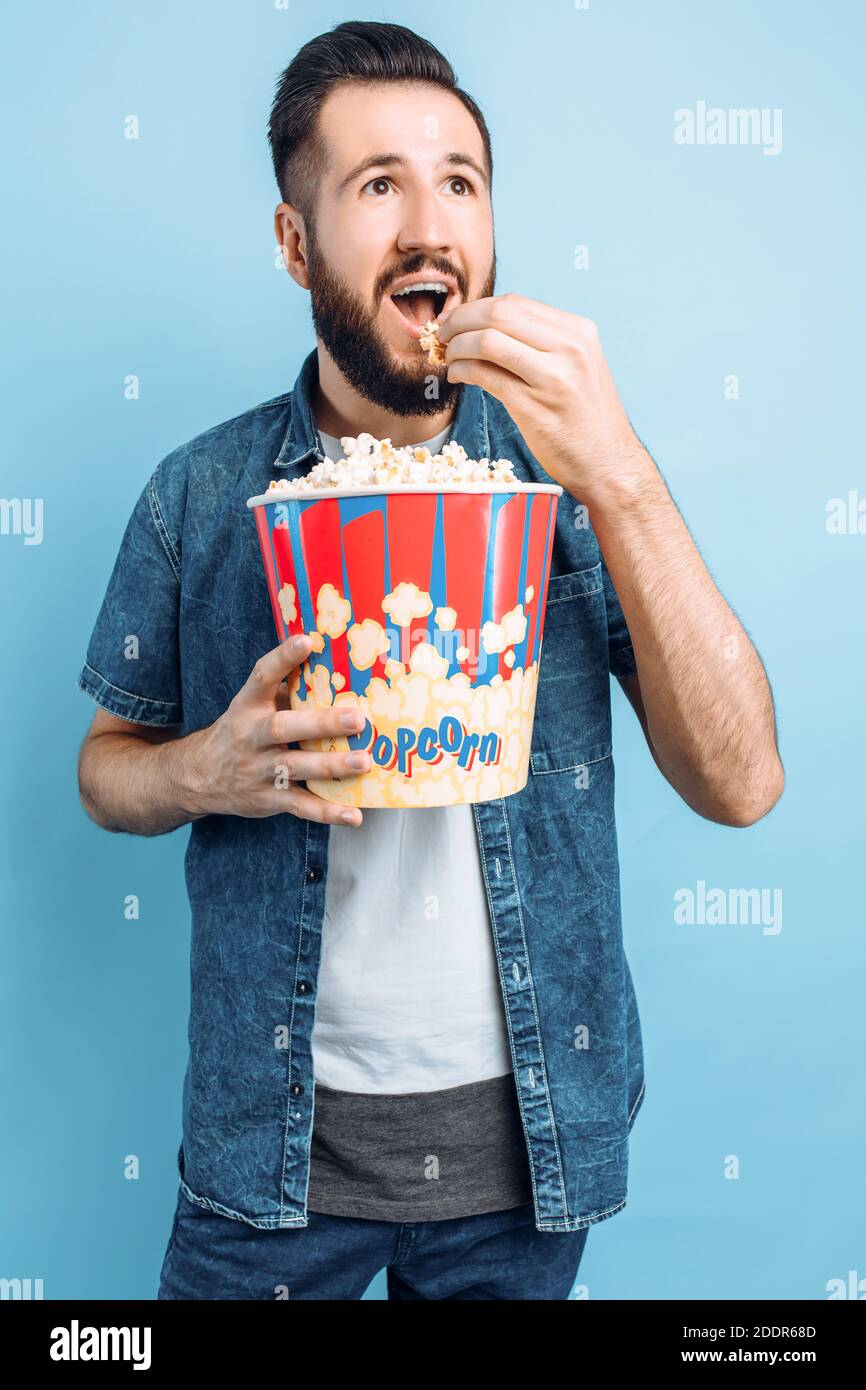 Felice uomo bearded tenendo un secchio di popcorn, trascorrere una serata in un ambiente accogliente casa, giovane ragazzo emotivo guardando un film, su un isolato bl Foto Stock