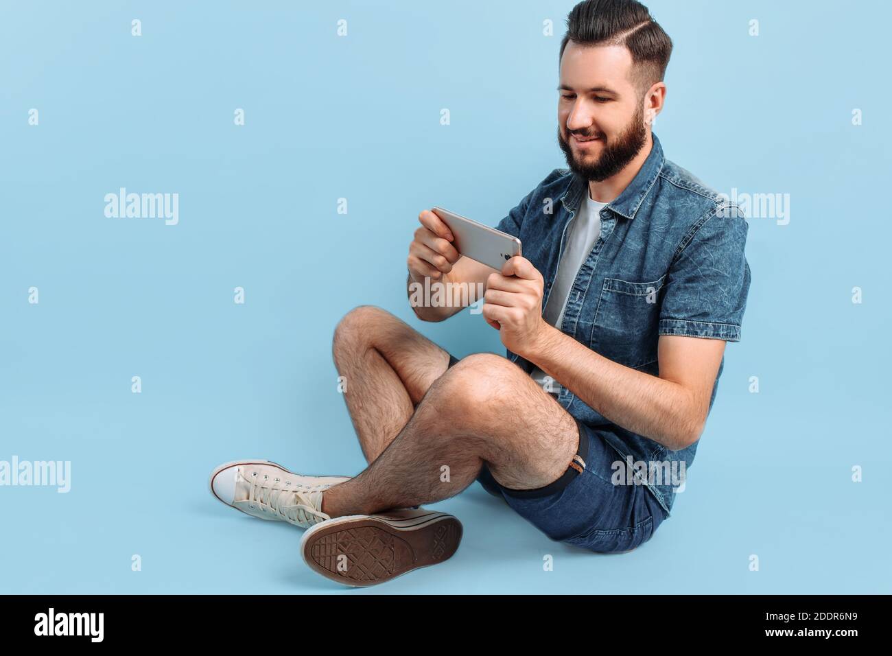 Un uomo con un'elegante bearded, in abiti casual, tiene uno smartphone nelle mani e guarda lo schermo del telefono seduto su uno sfondo blu Foto Stock