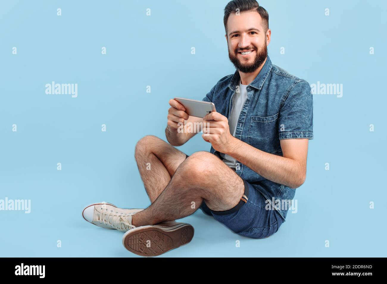 Un uomo con un'elegante bearded, in abiti casual, tiene uno smartphone nelle mani e guarda lo schermo del telefono seduto su uno sfondo blu Foto Stock