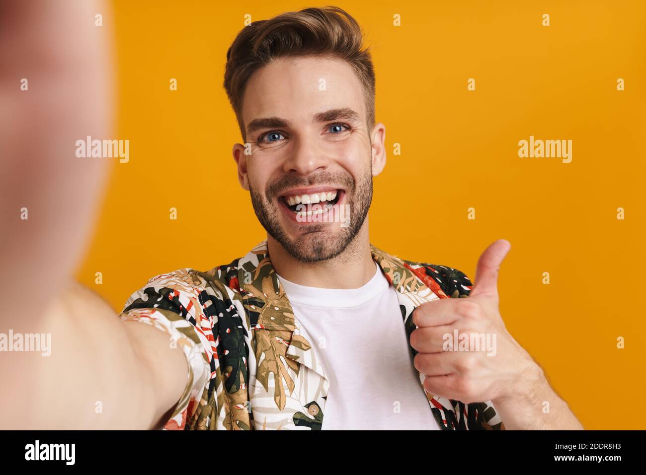 Allegro bel ragazzo gesturing pollice in su mentre scattano foto selfie isolato su sfondo giallo Foto Stock