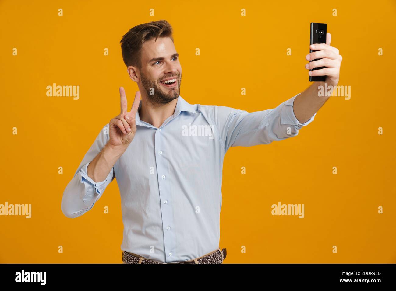 Foto di un uomo sorridente che gesturava il segno della pace mentre prendeva selfie sul cellulare isolato su sfondo giallo Foto Stock