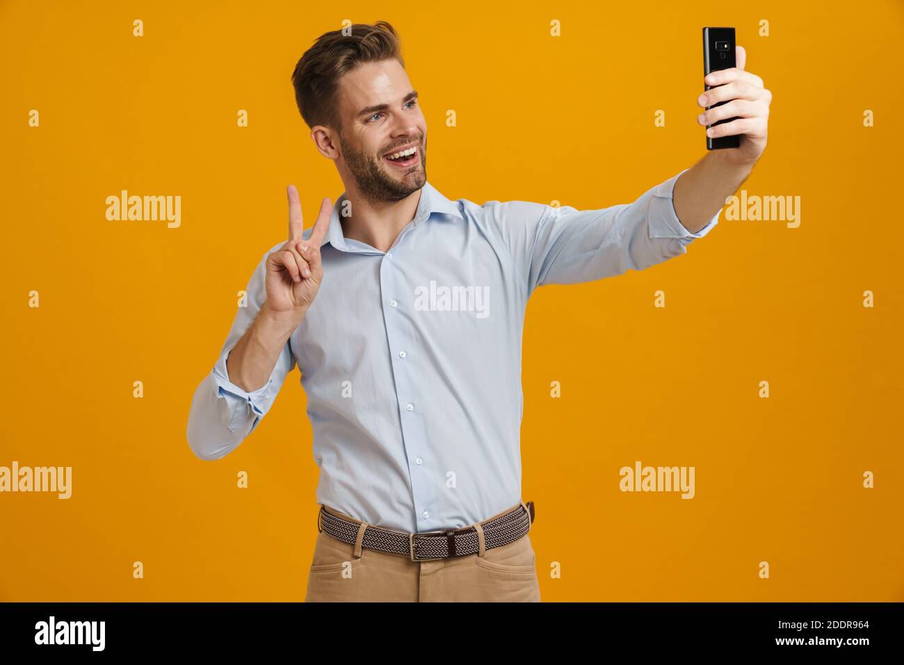 Foto di un uomo sorridente che gesturava il segno della pace mentre prendeva selfie sul cellulare isolato su sfondo giallo Foto Stock