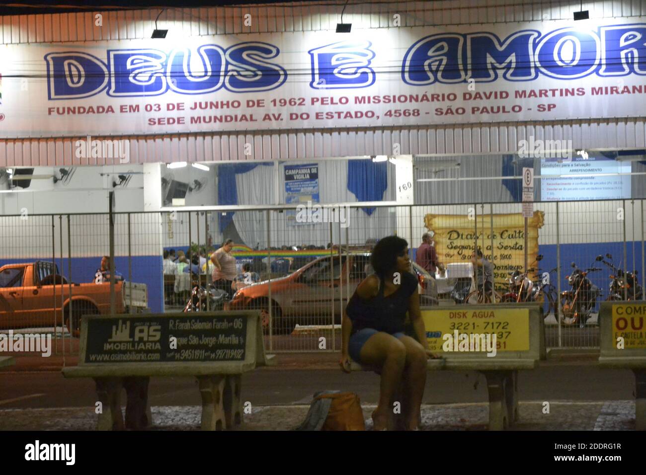 Giovane donna seduta su una panchina alla fermata dell'autobus, senza maschera, davanti alla pandemia del Covid 19, dietro una chiesa evangelica con un segno con il testo Foto Stock