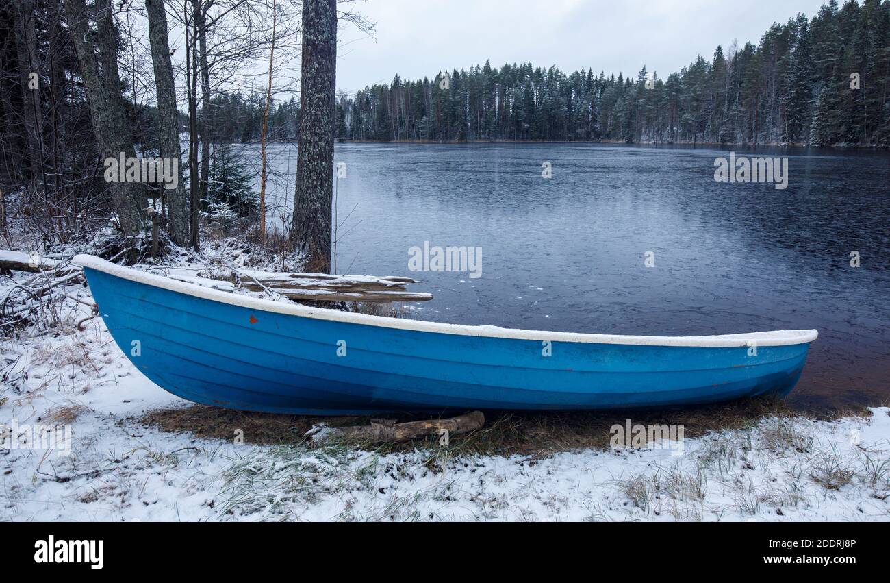 Una barca a remi in fibra di vetro blu affondata a riva di un lago coperto di sci di ghiaccio a Winter , Finlandia Foto Stock