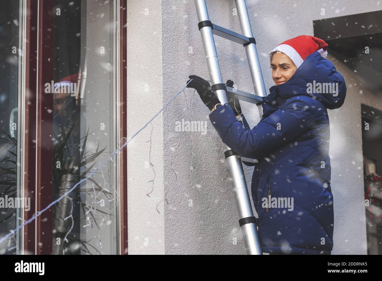 donna con cappello santa in piedi su scala e decorazione casa esterno con luci esterne stringa di natale. nevicare Foto Stock