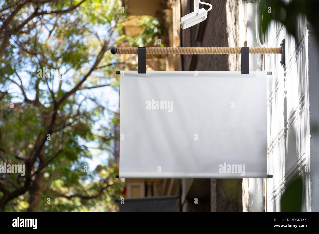 Cartello bianco appeso sulla facciata di un edificio all'ingresso di un negozio, con alberi sullo sfondo. Vuoto per messaggio personalizzato Foto Stock