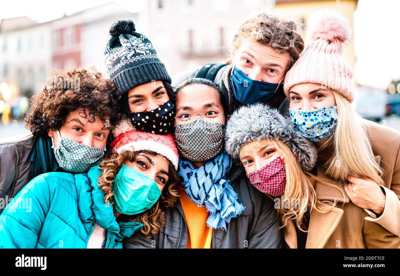 Amici multirazziali che prendono selfie indossando maschera facciale e vestiti invernali - nuovo concetto di stile di vita normale con i giovani che si divertono insieme all'esterno Foto Stock