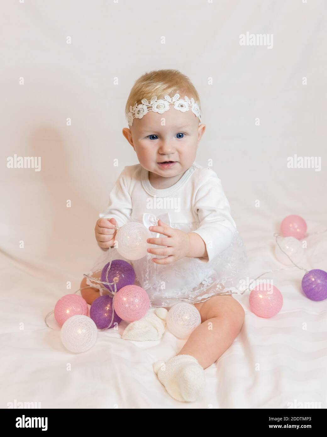 Una piccola ragazza di otto mesi in un abito bianco intelligente con palloncini garland nelle sue mani sorride. Sulla testa di un bendaggio bianco di Lacy Foto Stock