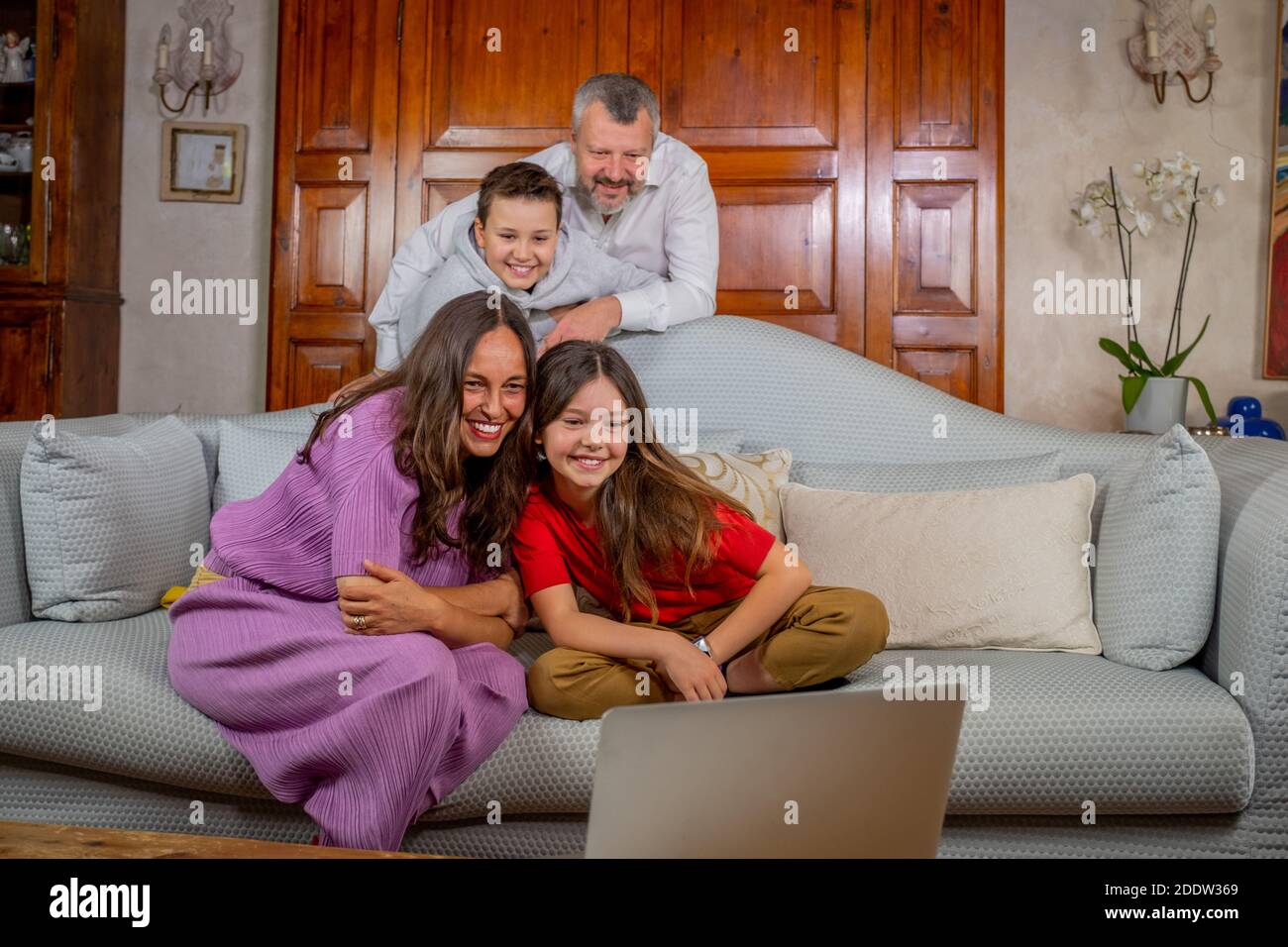 Felice famiglia che ha una videochiamata con gli amici, salutando lo schermo del portatile - Padre, madre, figlia e figlio sul divano a casa facendo una video chat w Foto Stock