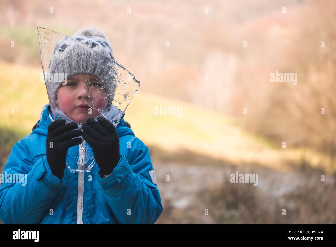 Ragazzo di cinque anni che tiene un piccolo pezzo di ghiaccio fondato nella foresta. Guardando attraverso iceberg alla fotocamera. Spazio per il testo. Concetto di arrivo dell'inverno. Foto Stock