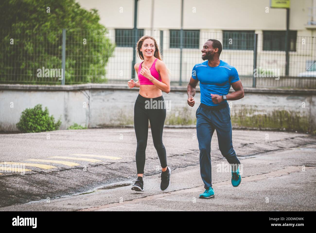 Giovane coppia sorridente l'un l'altro mentre si fa jogging sulla strada, fuori per correre. Afro americano uomo e bella ragazza bionda in sportswear che corre attraverso Foto Stock