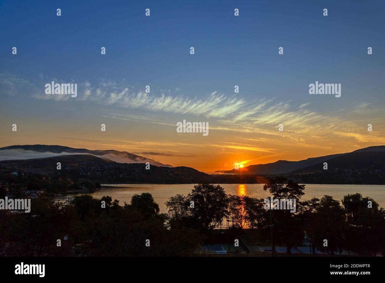 Alba su montagne e lago, vicino alla città costiera. Foschia di nebbia sulle montagne, il riflesso del sole nell'acqua, sagome degli alberi i Foto Stock