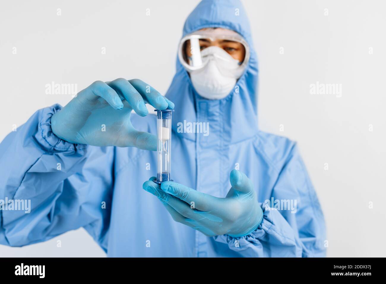 Medico maschile in un vestito protettivo e una maschera medica sul viso, guardando una clessidra con sabbia che scorre verso il basso, tempo limitato Foto Stock