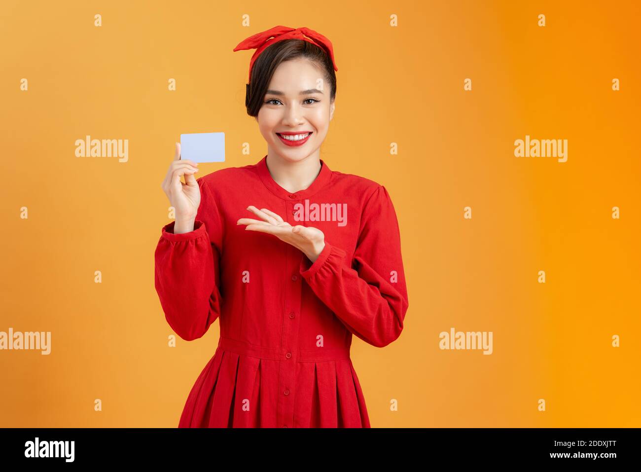 Giovane donna allegra in abito rosso che tiene la carta di credito della banca isolato su sfondo arancione Foto Stock