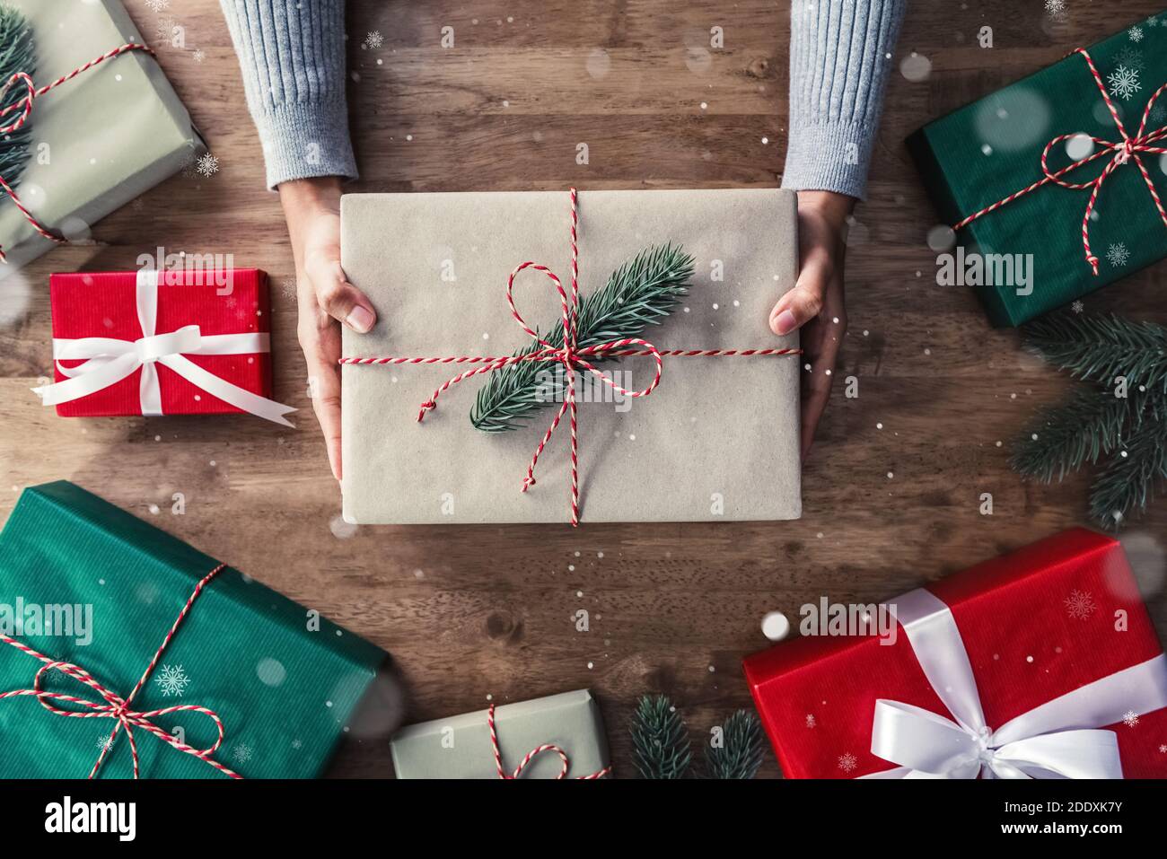 Le mani della donna che danno il regalo di Natale sul bordo del tavolo di legno con gli articoli e le scatole decoranti, preparati per celebrare la stagione festiva di festa, vista dall'alto Foto Stock