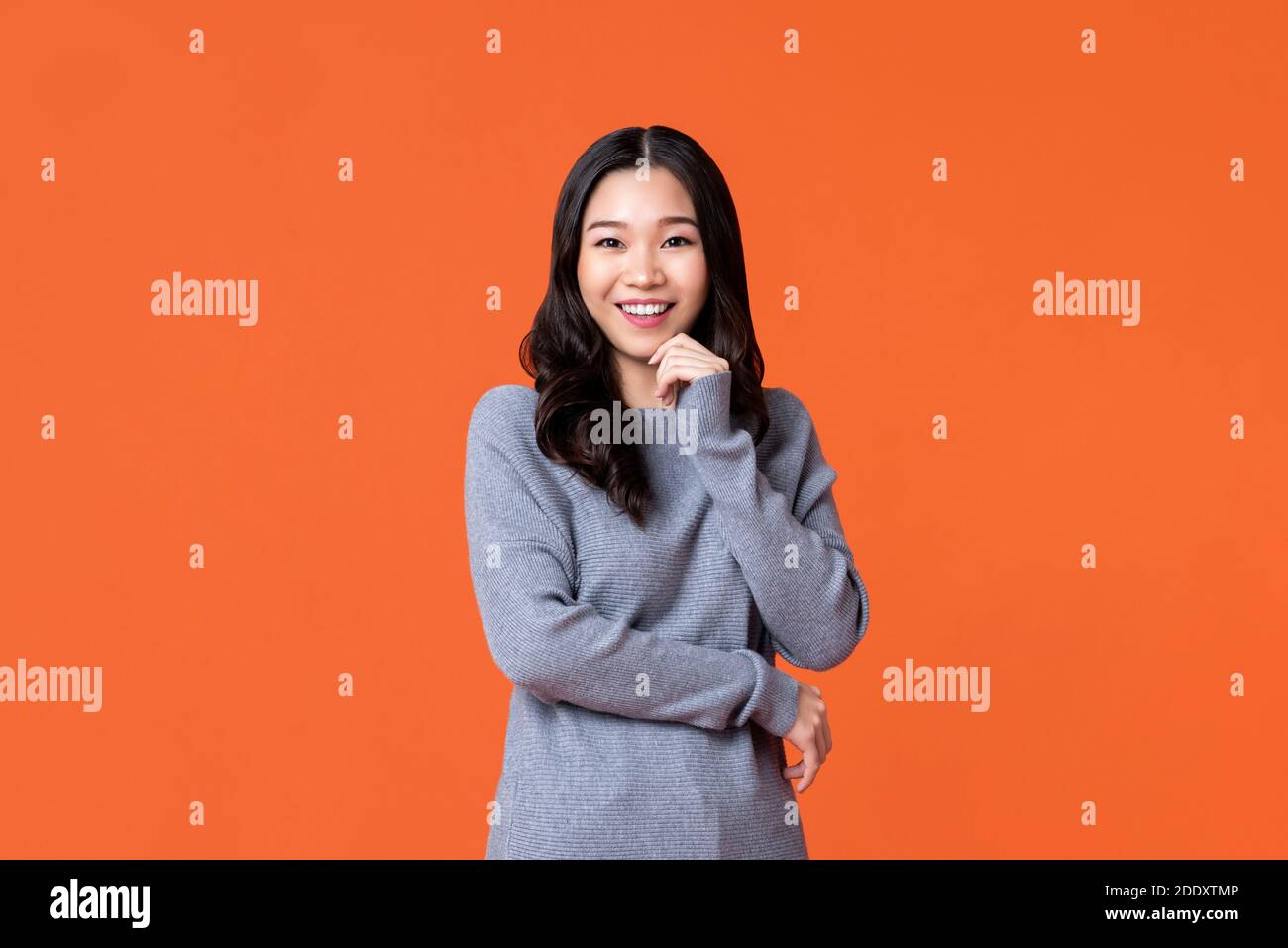 Giovane donna asiatica felice sorridente con la mano sul mento isolato su sfondo arancione studio Foto Stock