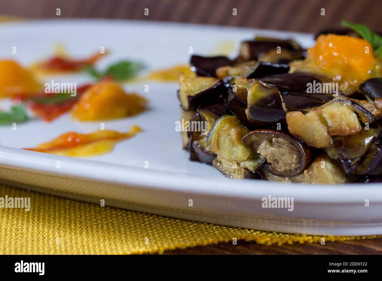 Piatto di melanzane a dadini su una crema di pomodori ciliegini gialli e rossi in un piatto bianco. Legno di fondo. Messa a fuoco selettiva. Foto Stock
