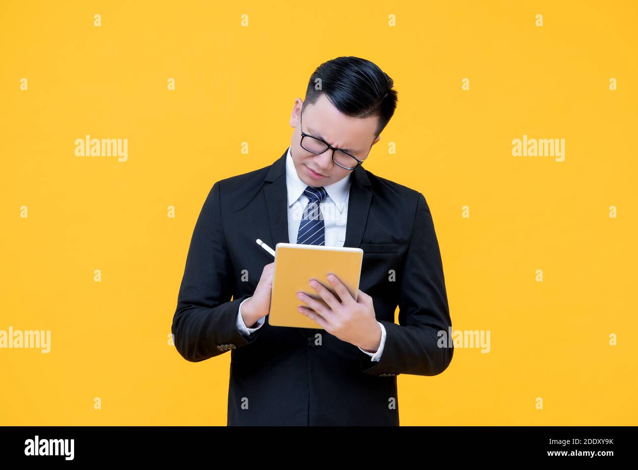 Uomo d'affari asiatico serio in tuta formale prendendo nota sul digitale computer tablet isolato su sfondo giallo Foto Stock